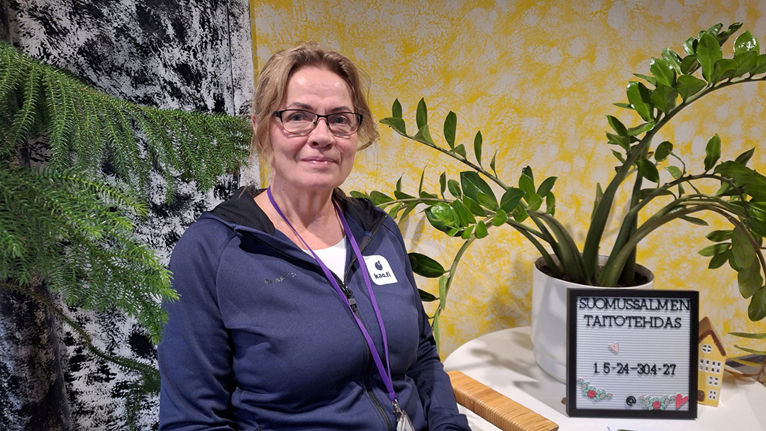 Silämälaispäinen henkilö puolilähikuvassa istuu tuolilla, takana näkyy kaksi viherkasvia. Oikealla olevalla pöydällä on Taitotehtaan kyltti. 