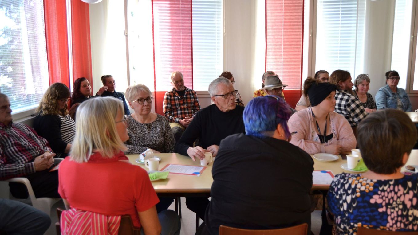 Kyläparlamentin osallistujia istuu pitkän pöydän äärellä, kolme on selin kameraan, pöydän takana ikkunoiden luona istuu kymmenkunta henkilöä. Ikkunoissa on punaiset ja valkoiset verhot.