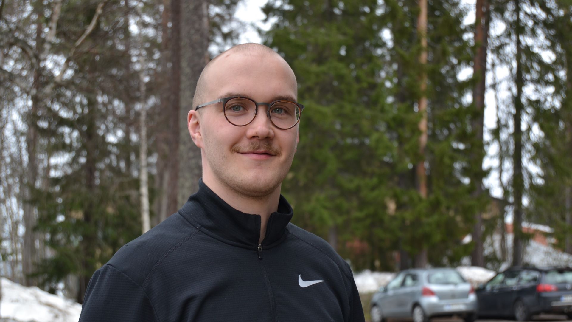 Hymyilevä silmälasipäinen henkilö puolilähikuvassa seisoo ulkona, taustalla kuusikkoista metsää ja vähän lumikasoja.