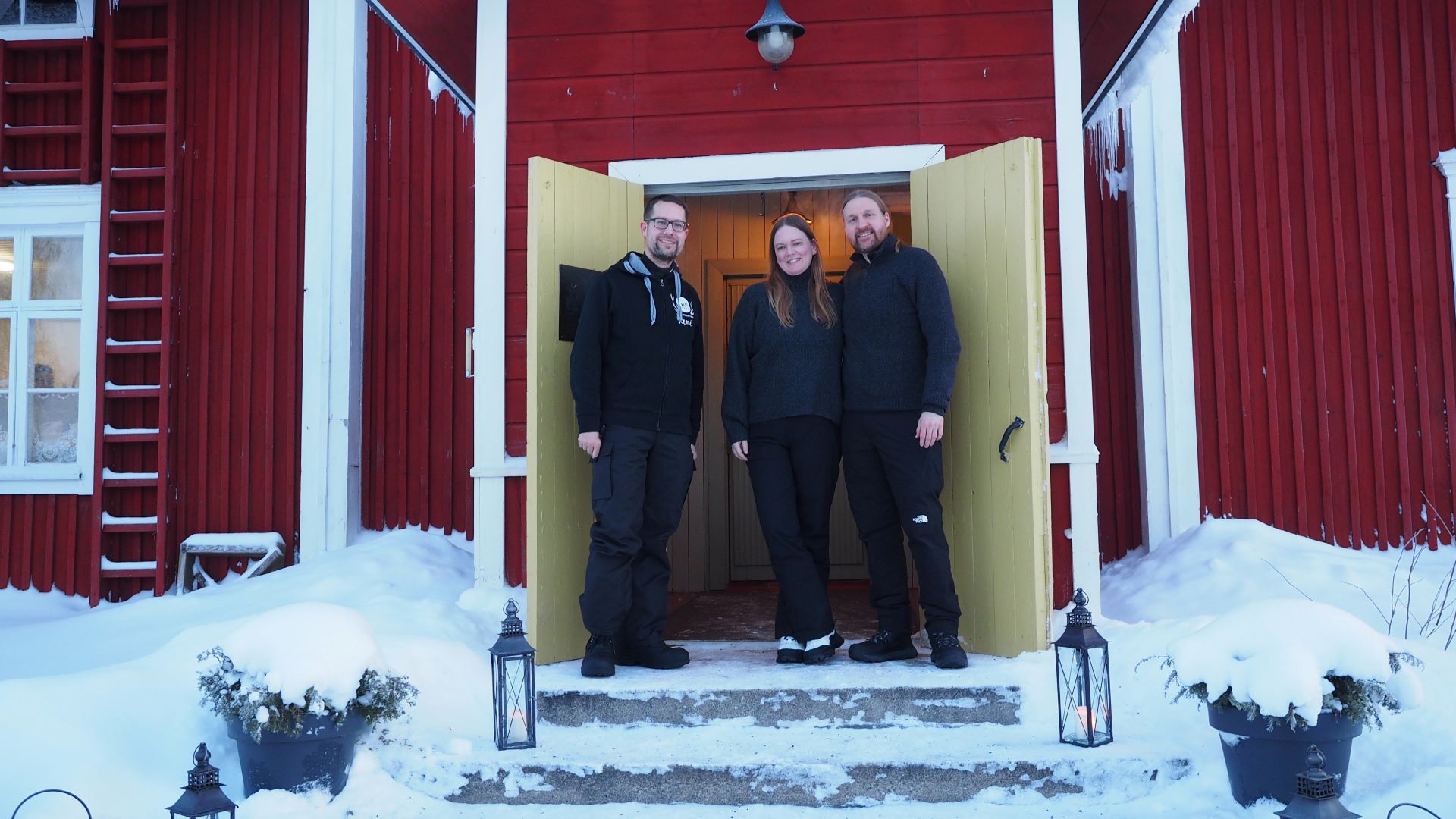 Kolme henkilöä seisoo vanhan punaisen puutalon oviaukossa.