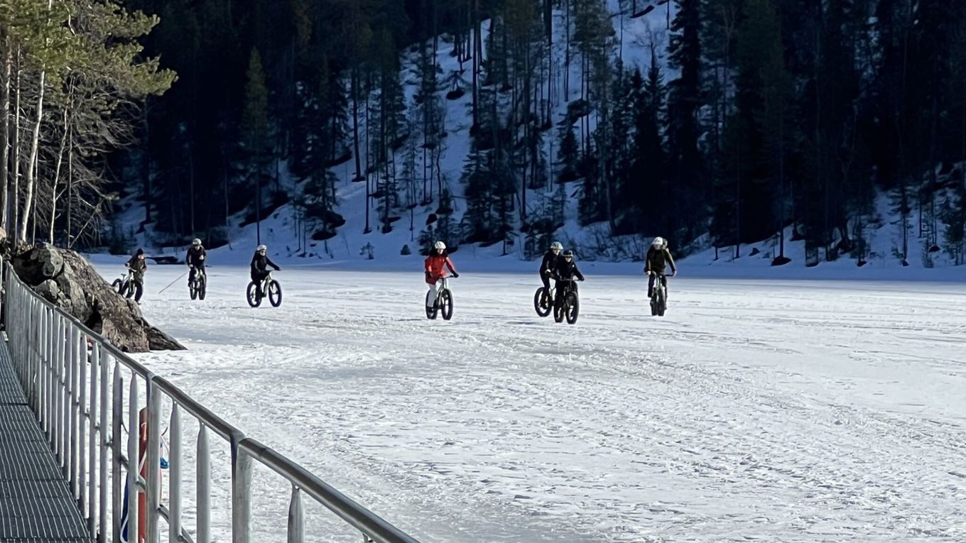 7 läskipyöräilijää jään päällä, taustalla jyrkkä metsäinen rinne ja etualalla metallista kaidetta.