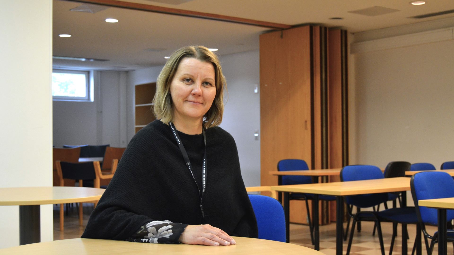 Hymyilevä, tummaan puseroon pukeutunut henkilö istuu pöydän äärellä. Toinen käsi lepää pöydällä. Hänen takanaan näkyy luentosalin pöytiä ja sinisellä kankaalla verhoiltuja tuoleja. Taustalla näkyy toinen tila, ja avoinna olevan tilanjakaja. 