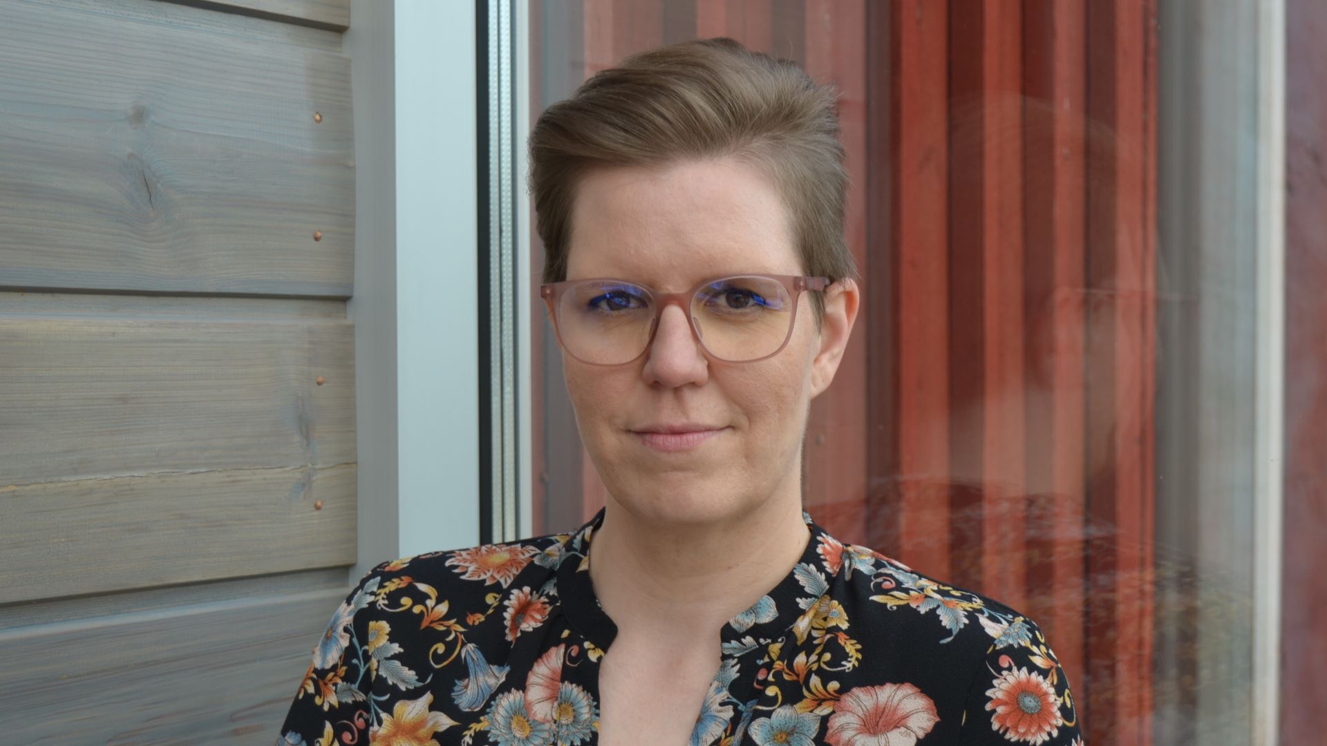 Lähikukuvassa lyhyttukkainen silmälasipäinen ja kirjavaan puseroon pukeutunut nainen. 