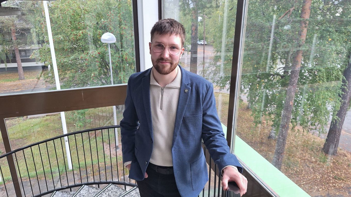 Puolilähikuvassa siniseen pikkutakkiin ja vaaleaan paitaan pukeutunut tummatukkainen ja silmälaispäinen mies seisoo portaikossa, takanaan isot ikkunat.