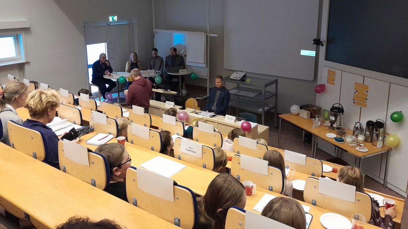 Kokouspaikkaa kuvattu takaa ja ylhäältä, näkyy ihmisiä istumassa paikoillaan, etualalla on viisi aikuista henkilö.