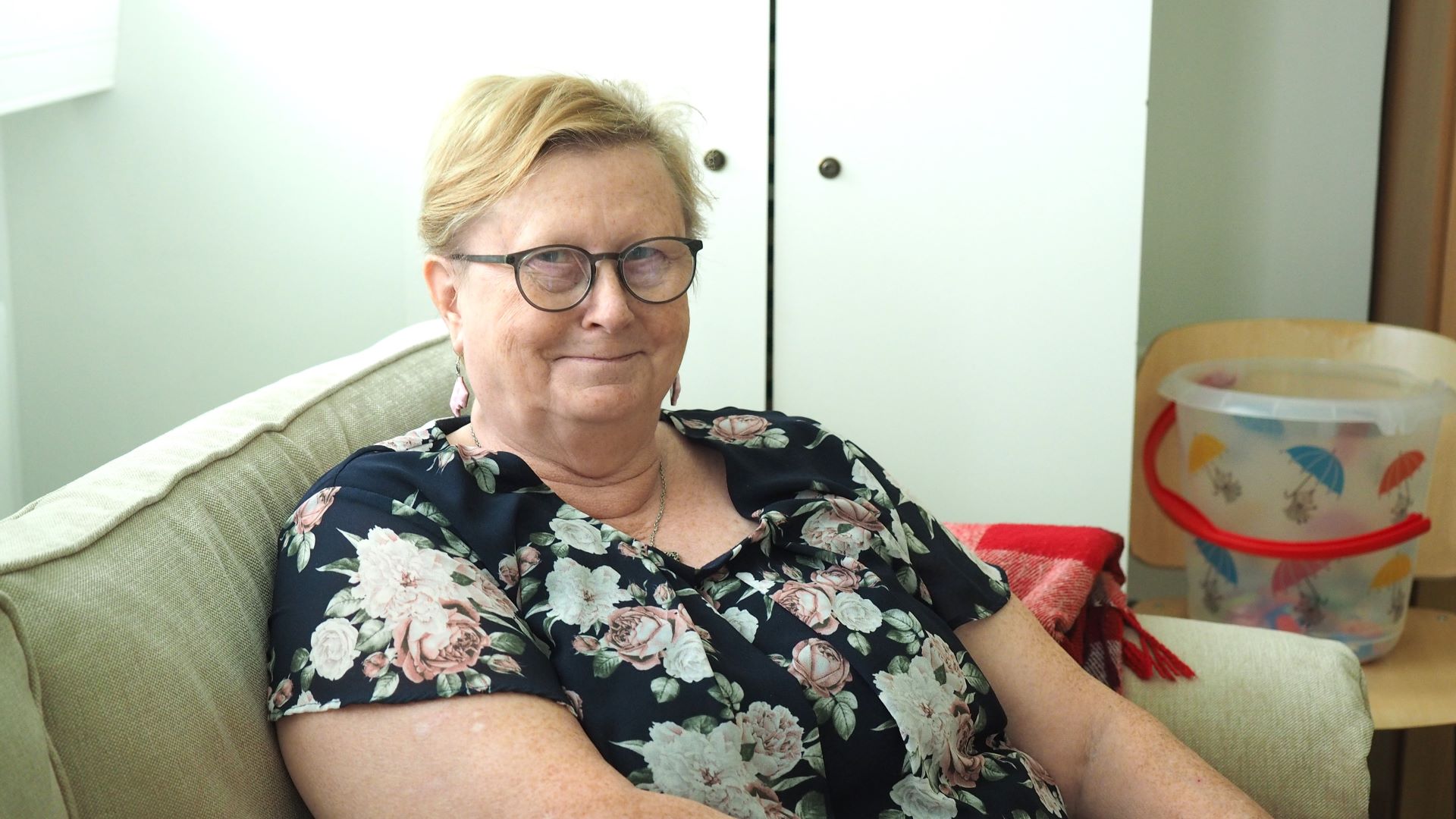 Puolilähikuva vaalealla sohvalla istuvasta hymyilevästä henkilöstä. Taustalla vaaleat kaapinovet, pienellä pöydällä värikäs ämpäri.