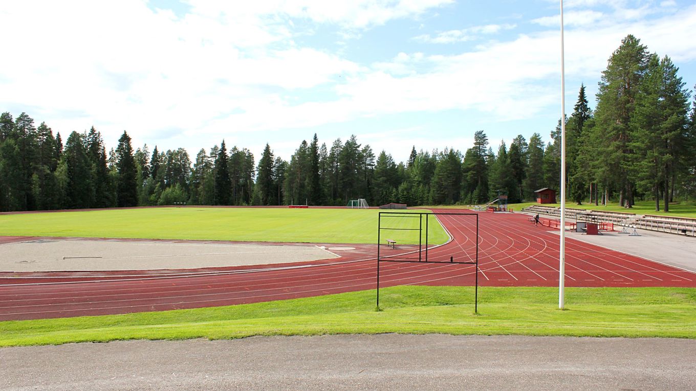 Kesällä kuvattu metsän ympäröimä urheilukenttä, juoksuratojen takana nurmikenttä.