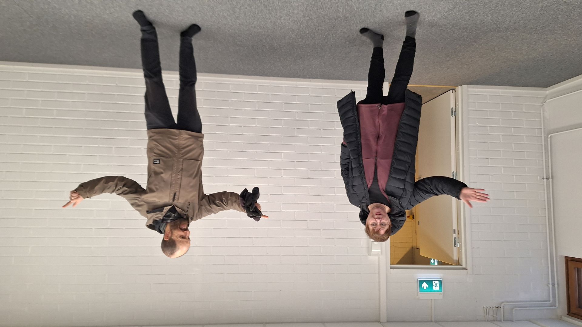 Tyhjässä valkeaseinäisessä huoneessa kaksi henkilöä seisomassa, he osoittavat käsillään sivulle.