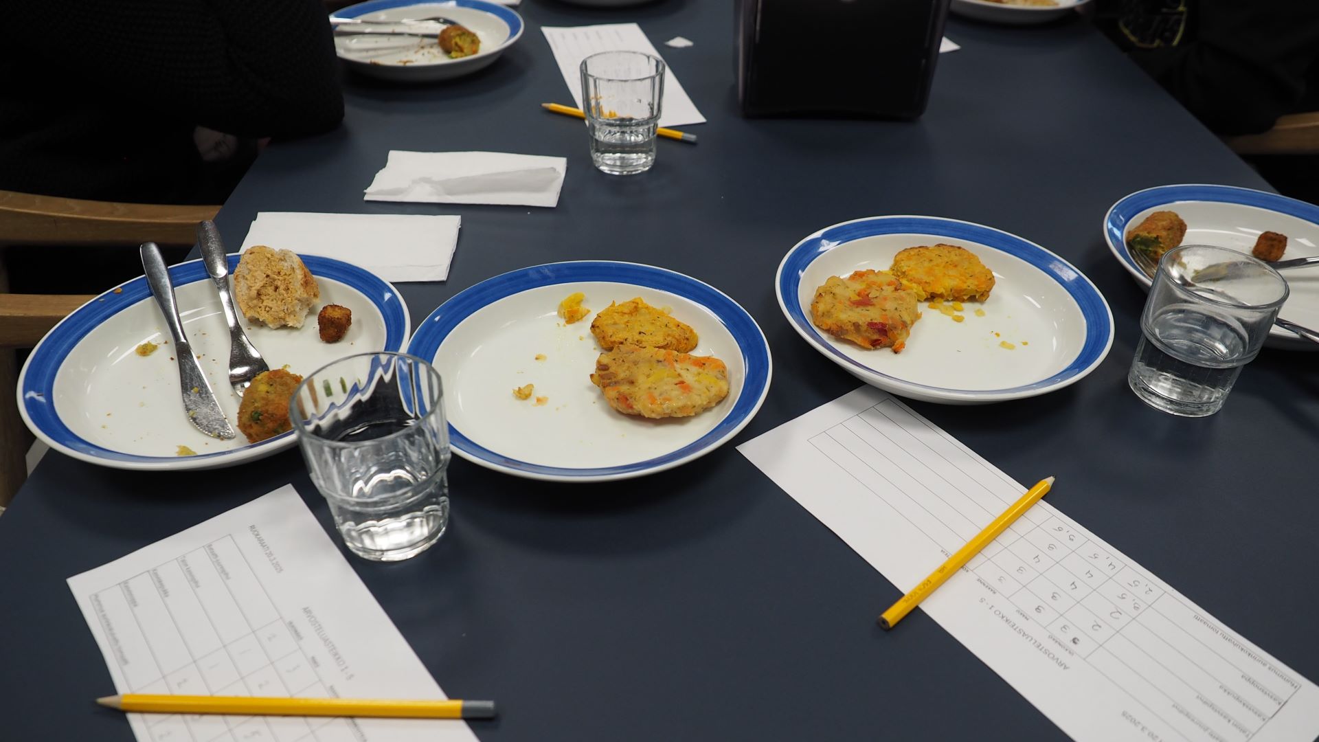 pöydällä on useita lautasia, jolla osittain syötyjä kasvispihvejä, pöydällä on myös muutama juomalasi, joissa vettä, lisäksi arvostelulomakkeita ja lyijykyniä.
