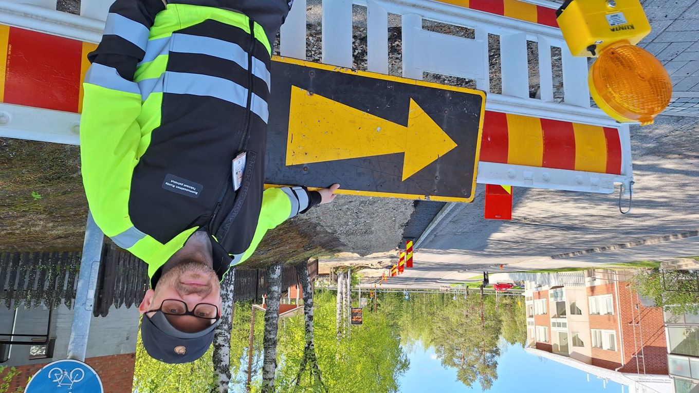 Puolilähikuvassa hymyilevä, lippalakkiin ja huomiovaatteisiin pukeutunut silmälaispäinen mies seisoo tilapäisen esteaidan edessä. Takana näkyy katutyömaa.