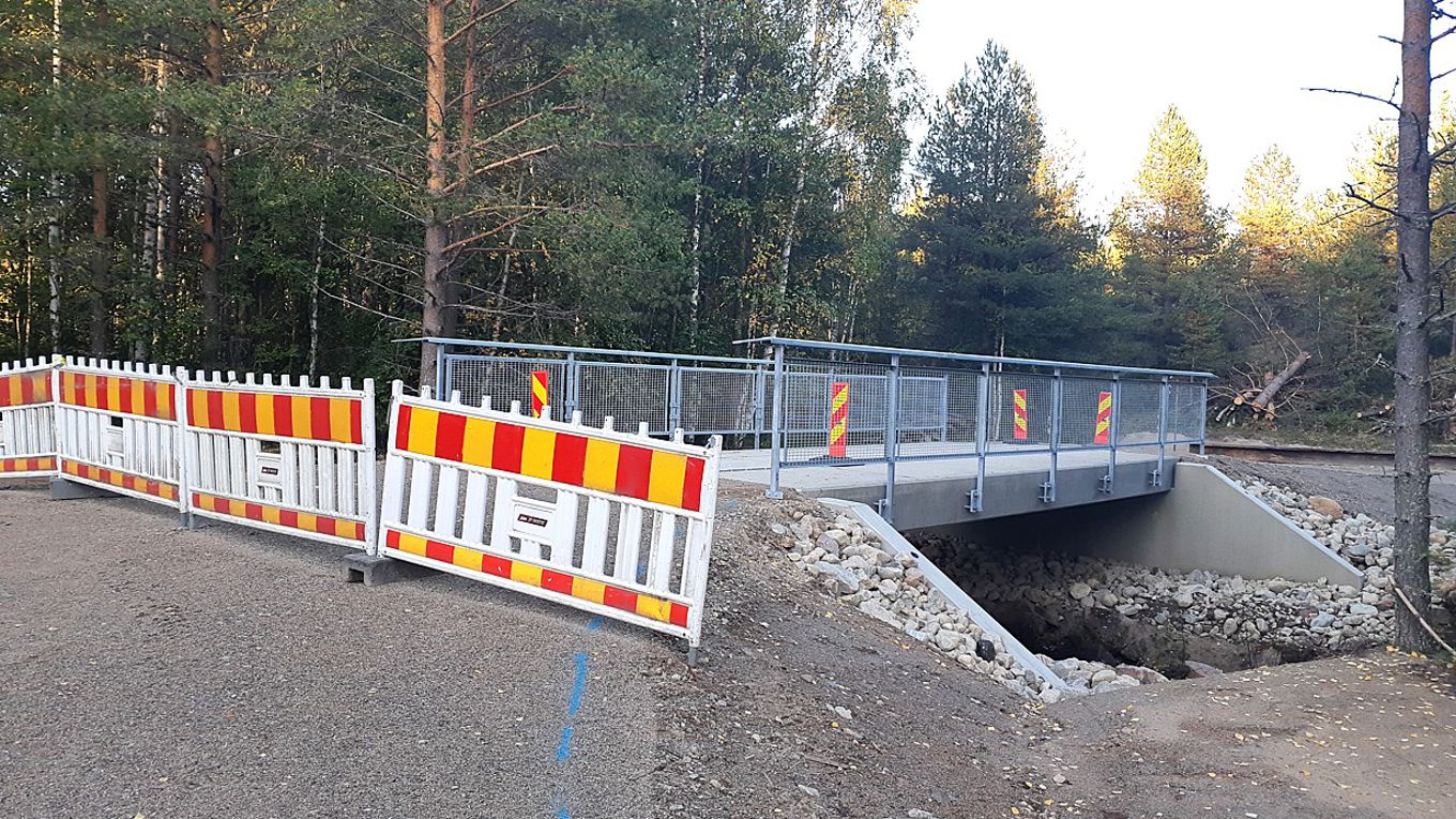 Etualalla on työmaa-aidoin suljettu betonirakenteinen yksinkertainen silta, jossa on metalliset, keskeneräisen näköiset kaiteet. Taustalla on metsää.
