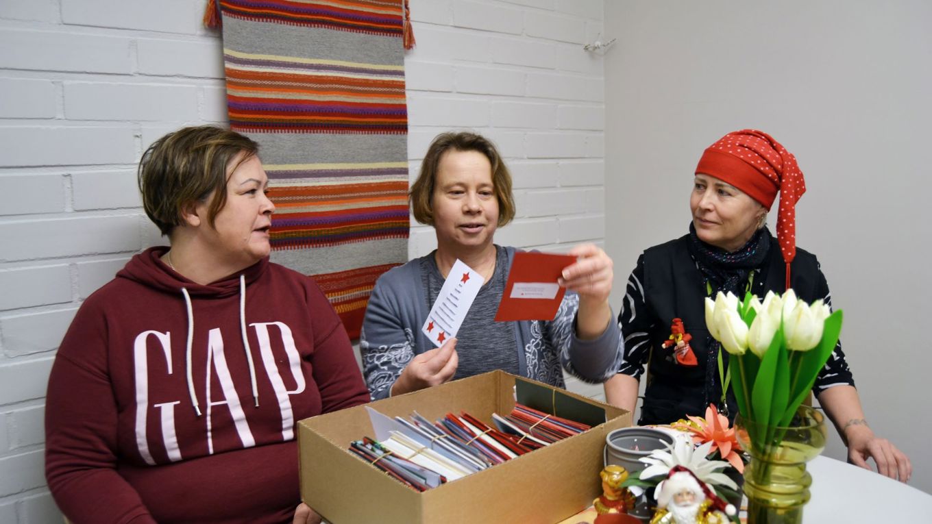 Kolme henkilöä istuu pöydän ääerellä, yksi pitelee kahta joulukorttia, pöydällä on laatikko, jossa paljon kortteja.