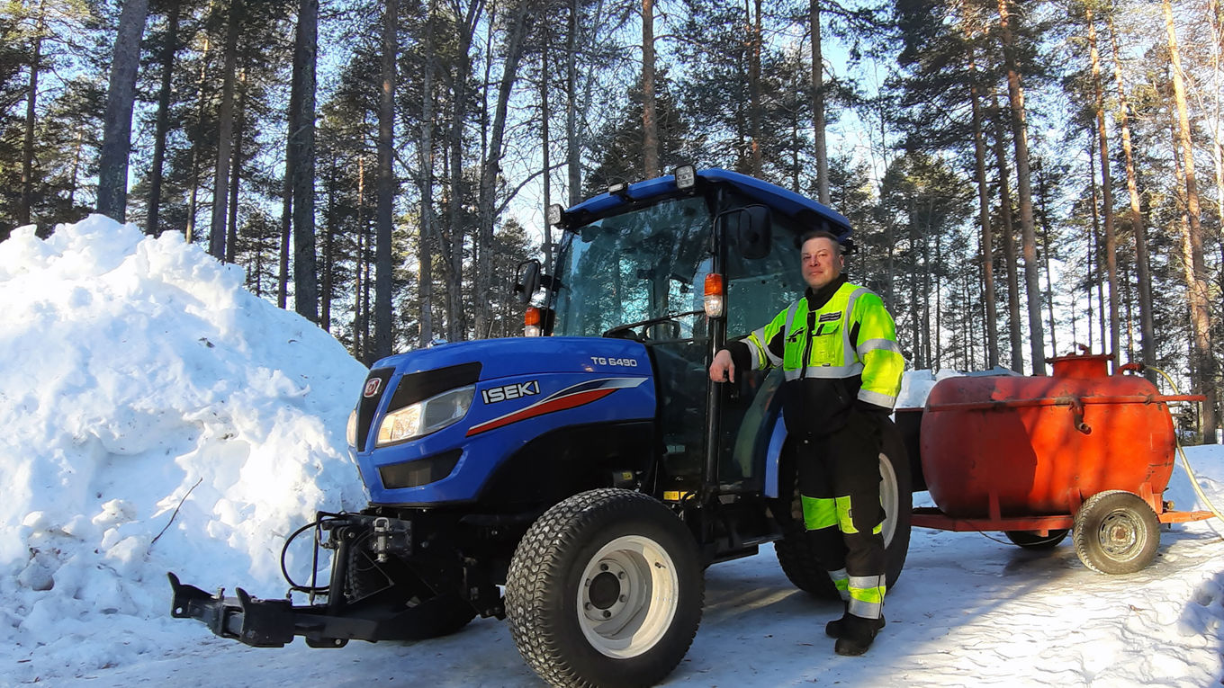 Työasuun pukeutunut henkilö poseeraa sinisen traktorin vierellä, traktorin perässä on punainen vesisäiliö pyörien päällä. Vasemmalla on iso lumikasa, taustalla mäntymetsää.