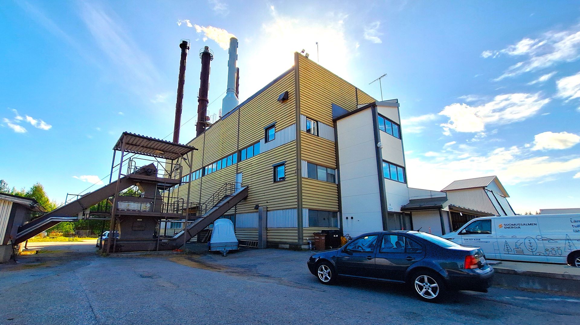 Lämpölaitos ulkoa kesällä kuvattuna, etualalla asvalttia ja pari autoa. 