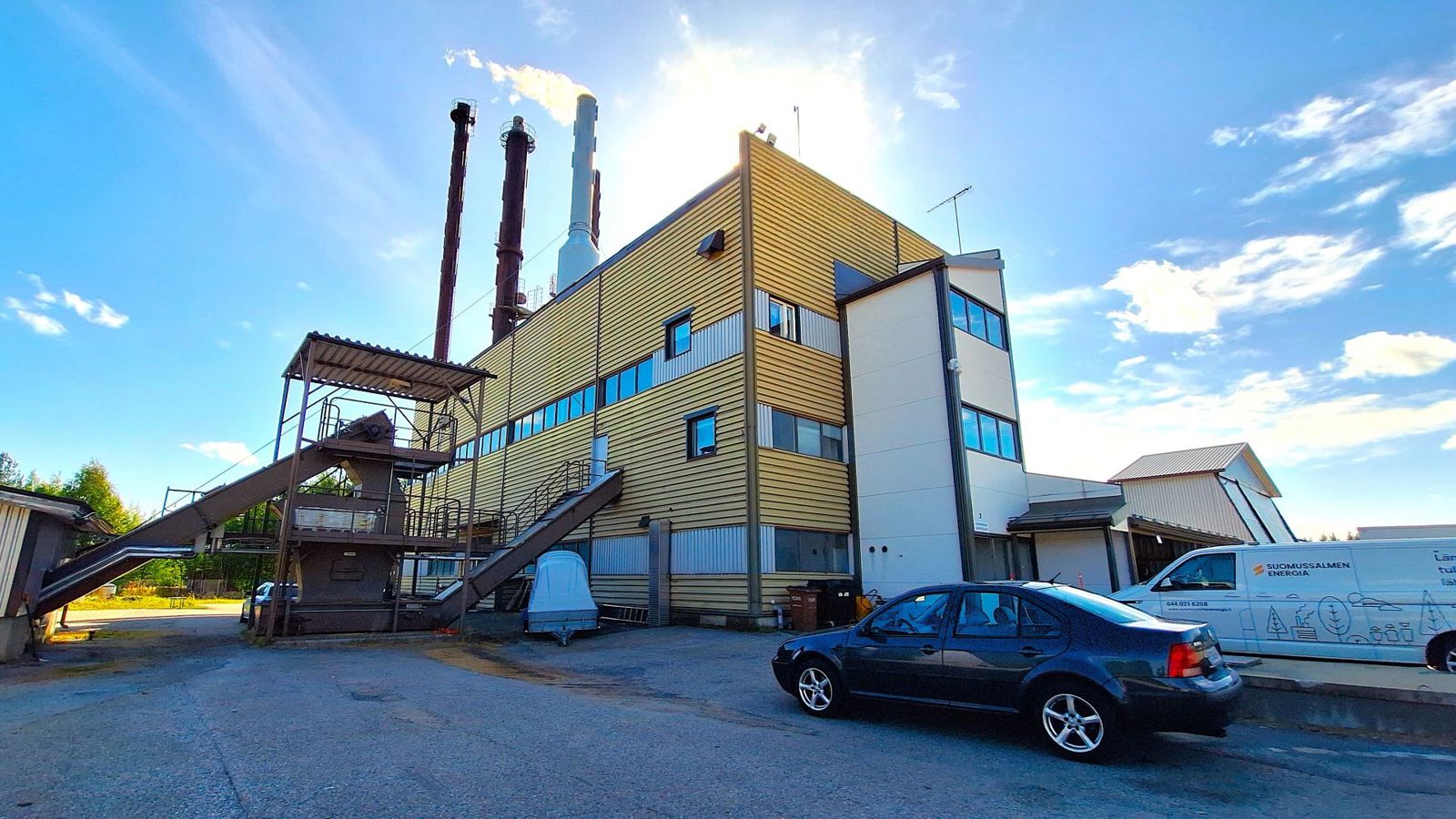 kaukolämpölaitos kuvattuna kesällä, etualalla asvalttia ja pari autoa, taivas on sininen, vähän valkoisia pilviä taivaalla, rakennuksen takaa aurongonvalo hohkaa valkoisena.