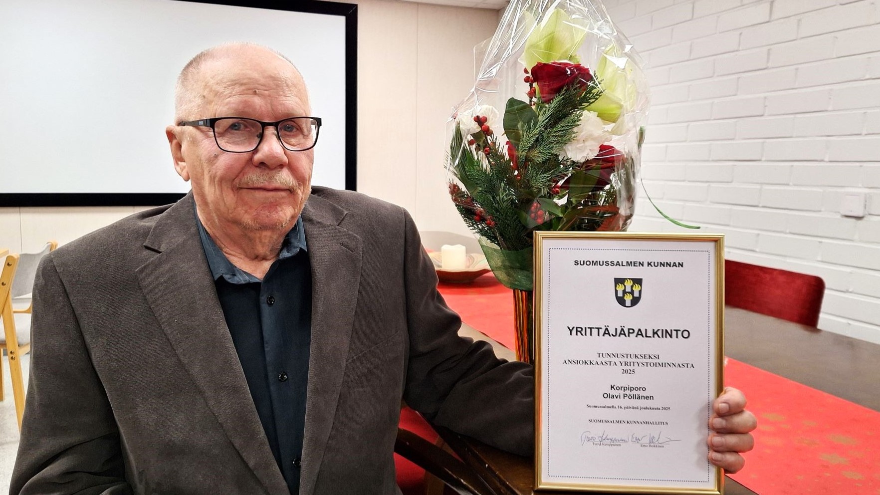 Henkilö lähikuvassa, istuu pöydän äärellä ja pitelee kehystettyä kunniakirjaa, pöydällä myös kaunis kukkakimppu maljakossa. 