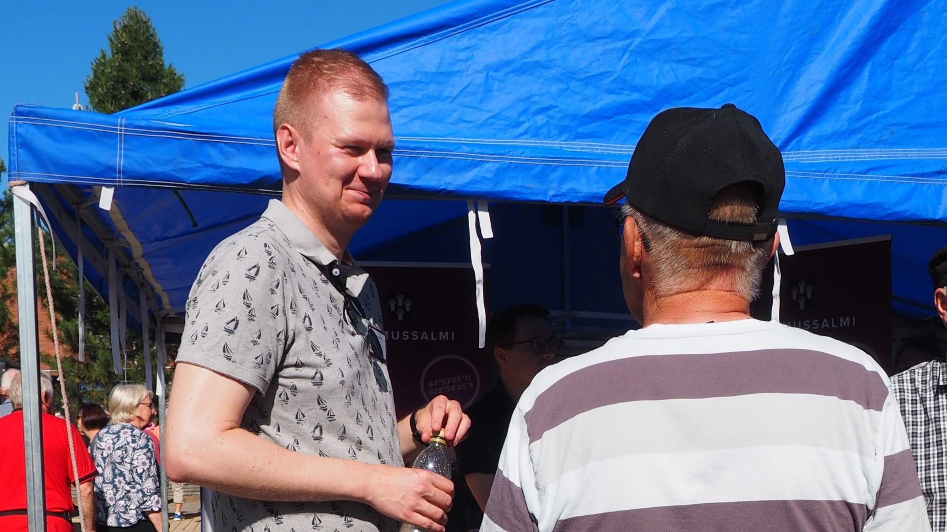 Hymyilevä, kirjavaan paitaan pukeutunut henkilö kasvot kameran suuntaan keskustelee selin kameraan olevan lippalakkisen henkilön kanssa, säätila näyttää kuumalta, aurinko paistaa, taustalla näkyy ihmisiä ja ylimpänä sinistä katoskangasta.