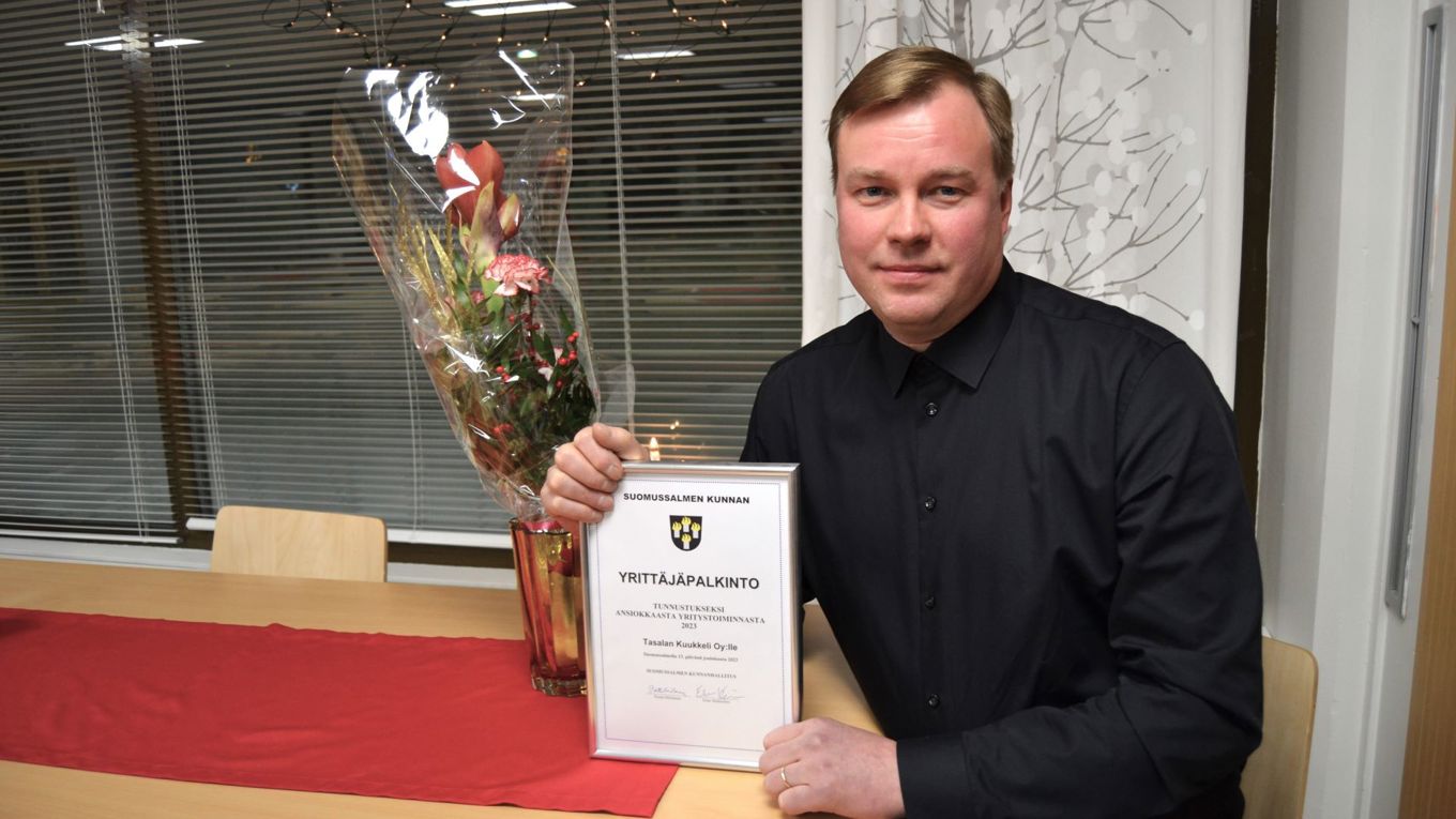 Henkilö istuu pöydän vierellä ja pitelee kehystettyä kunniakirjaa, vierellään pöydällä on kukkamaljakko, jossa kukkia. Pöydällä on punainen liina.