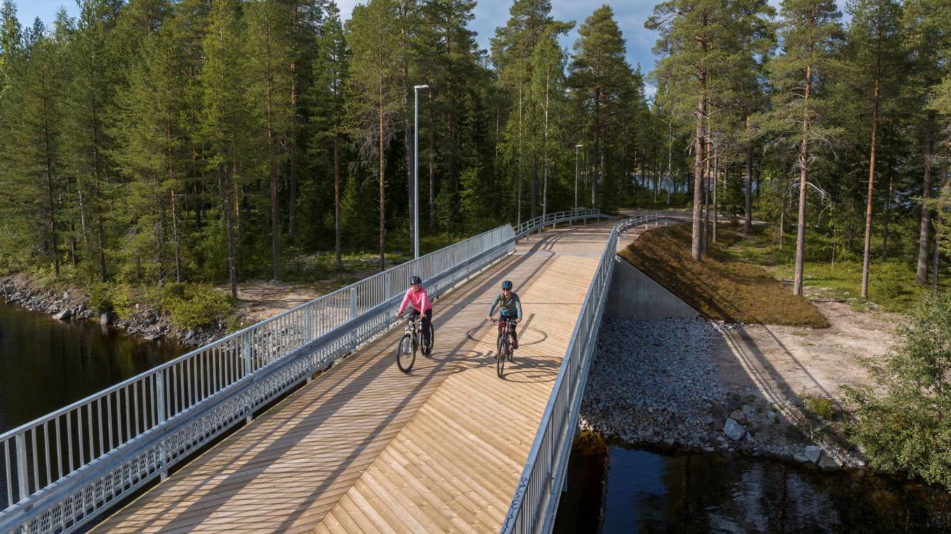 Kaksi pyöräilijää ylittää uutta puukantista siltaa, sillassa on metalliset kaiteet. Taustalla puustoa, muun muassa lehdessä olevia koivuja. Sillan alla näkyy sinistä vettä.