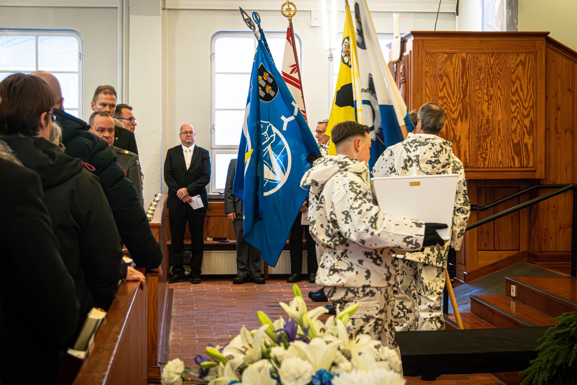 Kirkossa, vasemmalla seisoo joitakin tummiin pukeutuneita henkilöitä. Talvipukuinen sotilas kantaa sylissään valkoista pienehköä arkkua. Takana on asetettu lippuja telineeseen. Etualalla on kukkalaite, jossa valkoisia kukkia. 