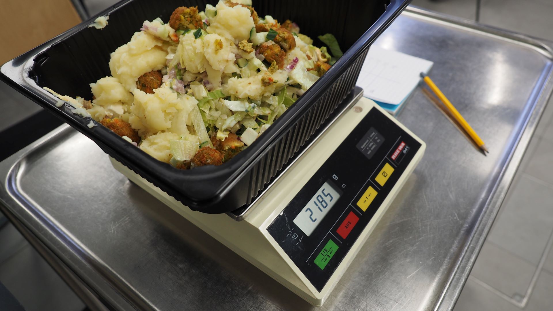 Digitaalisen vaa'an päällä on astia, ja siinä ruokajätettä. Vaa'an vieressä näkyy paperia ja kynä.