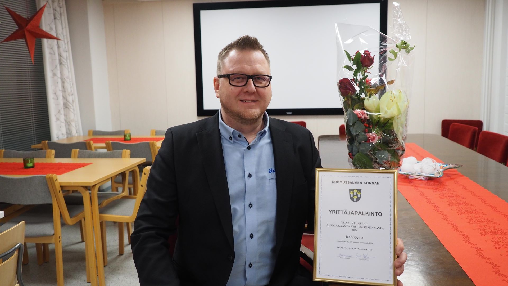 Hymyilevä mieshenkilö istuu pöydän äärellä, käsi pitelee kehystettyä palkintoplakaattia, takana pöydällä näkyy kukkakimppu.